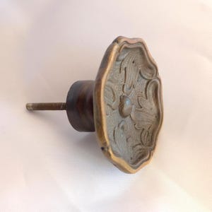May include: A round metal cabinet knob with a dark bronze finish and a scalloped edge. The face of the knob features a raised, symmetrical floral relief pattern with a central circular detail. A threaded metal bolt extends from the back.