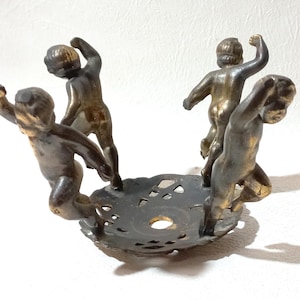 Könnte beinhalten: Antiker bronzefarbener Kerzenhalter mit vier cherubinen Figuren um einen dekorativen, durchbrochenen Sockel. Die Cherubim befinden sich in verschiedenen Posen, mit erhobenen Armen. Der Kerzenhalter ist ein dekorativer Artikel.