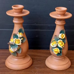 May include: Two terracotta candle holders. Each has a wide base, a central column, and a small, shallow cup at the top. The columns are decorated with yellow rose blossoms and green leaves. The candle holders are displayed on a wooden surface.