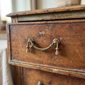 Puede incluir: Un tirador de cajón de latón con un asa curva de textura de cuerda y extremos decorativos en forma de borla, montado en un frente de cajón de madera marrón oscuro desgastada con veta visible y signos de antigüedad y desgaste.