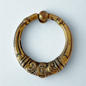 Puede incluir: Tirador de cajón de latón ornamentado con un asa de anillo y detalles decorativos. El tirador presenta un diseño circular con un pequeño pomo redondo para el montaje. El latón tiene un aspecto ligeramente envejecido, con acentos oscuros en las ranuras.