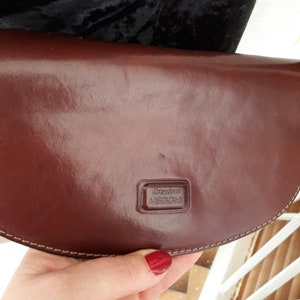 May include: A brown leather clutch with a curved shape. The clutch has a shiny finish and a stitched border. The brand name "Creazioni MEGGHI" is embossed on the front. The clutch is held by a person.