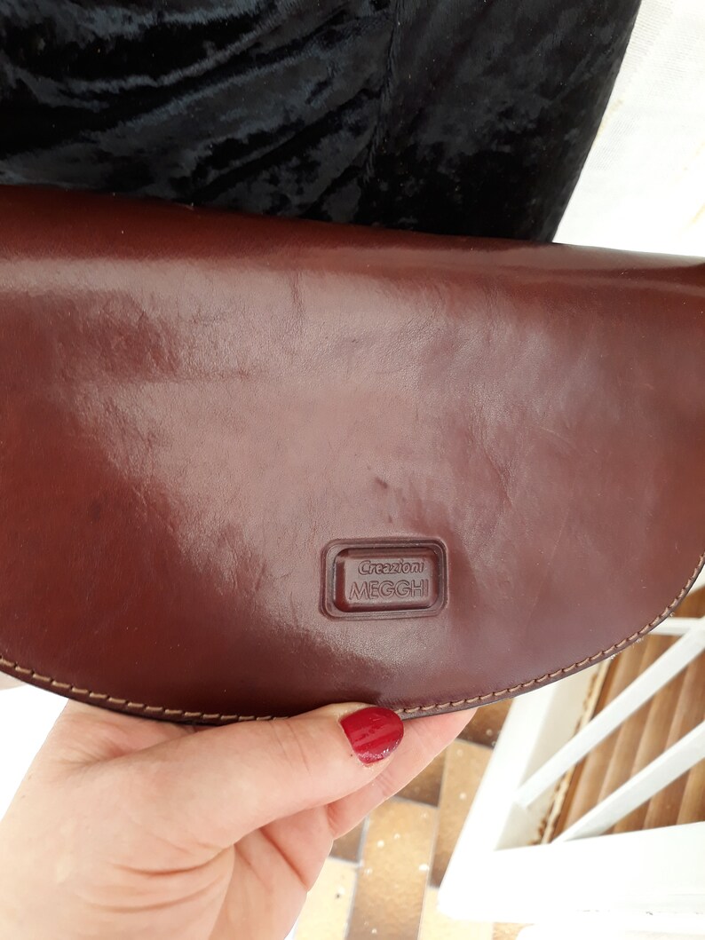 May include: A brown leather clutch with a curved shape. The clutch has a shiny finish and a stitched border. The brand name "Creazioni MEGGHI" is embossed on the front. The clutch is held by a person.