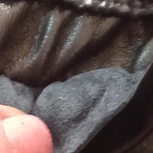 May include: Close-up of dark, textured fabric with visible stitching. The material is a deep black or charcoal colour, with a slightly glossy sheen. The image focuses on the texture and details of the material.
