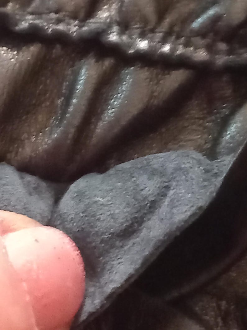 May include: Close-up of dark, textured fabric with visible stitching. The material is a deep black or charcoal colour, with a slightly glossy sheen. The image focuses on the texture and details of the material.