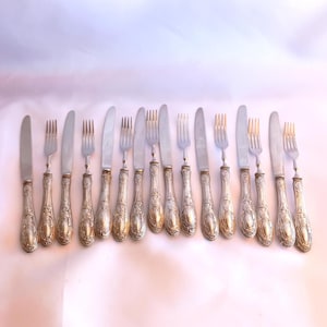 May include: A set of silver-toned flatware consisting of multiple knives and forks arranged in a row. Each piece features an ornate, embossed handle with intricate scrollwork and a polished metal finish on the blades and tines.