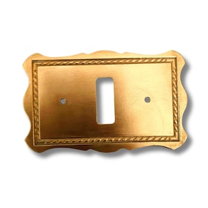 Puede incluir: Cubierta de interruptor de luz dorada, rectangular, con un borde decorativo en forma de cuerda. Tiene una abertura rectangular para el interruptor y dos pequeños orificios de montaje. Los bordes son festoneados.