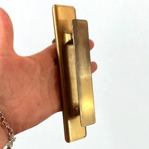 Peut inclure: Une poignée de porte vintage en laiton. Elle présente une plaque arrière rectangulaire et une longue poignée rectangulaire. Le laiton a une patine chaude et vieillie. La poignée est tenue dans une main.