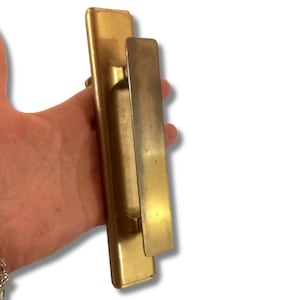 Puede incluir: Un tirador de puerta de latón antiguo con una base rectangular y una palanca larga y plana. El tirador tiene un tono dorado cálido y muestra signos de edad, lo que sugiere que es una antigüedad o una pieza de recuperación arquitectónica.