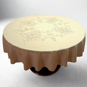 May include: A round, cream-colored tablecloth with an embroidered floral design. The tablecloth has a scalloped edge and is draped over a dark wooden table. The embroidered pattern features flowers and leaves in shades of yellow and green.