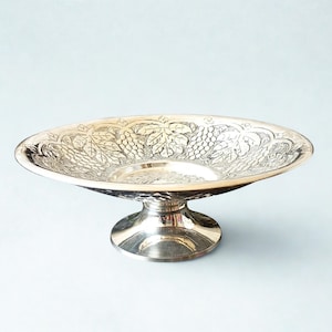Puede incluir: Cuenco con pedestal plateado de forma ovalada. El cuenco presenta un diseño en relieve de uvas y hojas alrededor del borde. La base es un pedestal corto y redondo.