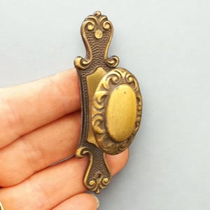 Puede incluir: Un tirador de puerta antiguo de latón con un pomo ovalado y detalles ornamentados. El tirador tiene una placa trasera rectangular texturizada y adornos decorativos. El latón tiene una pátina cálida y envejecida, que sugiere encanto vintage y diseño clásico.