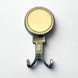 Puede incluir: Un perchero doble de estilo vintage con una parte superior circular y dos ganchos curvos. El gancho presenta un acabado de latón con detalles decorativos y una base de metal azul-gris oscuro. El diseño es simétrico.