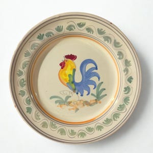 Peut inclure: Une assiette ronde en céramique avec un motif de coq peint à la main. Le coq est jaune, bleu et rouge, avec de l'herbe verte et un fond beige. L'assiette a un bord beige avec un motif de feuilles vertes et un accent orange.