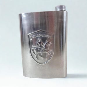 May include: A silver-colored flask with a screw-on cap. The flask features an embossed emblem of a person on horseback with the word "Запорожье" above. The flask is rectangular with rounded edges.