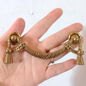 Puede incluir: Un tirador de cajón de latón con un asa en forma de cuerda y extremos decorativos de borla. El herraje presenta un diseño curvo con detalles intrincados, adecuado para la restauración de muebles o proyectos decorativos.