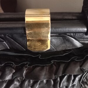 May include: Close-up of a black leather handbag with a gold clasp. The clasp is rectangular and features the Valentino Garavani logo. The bag has a textured, ruffled design.
