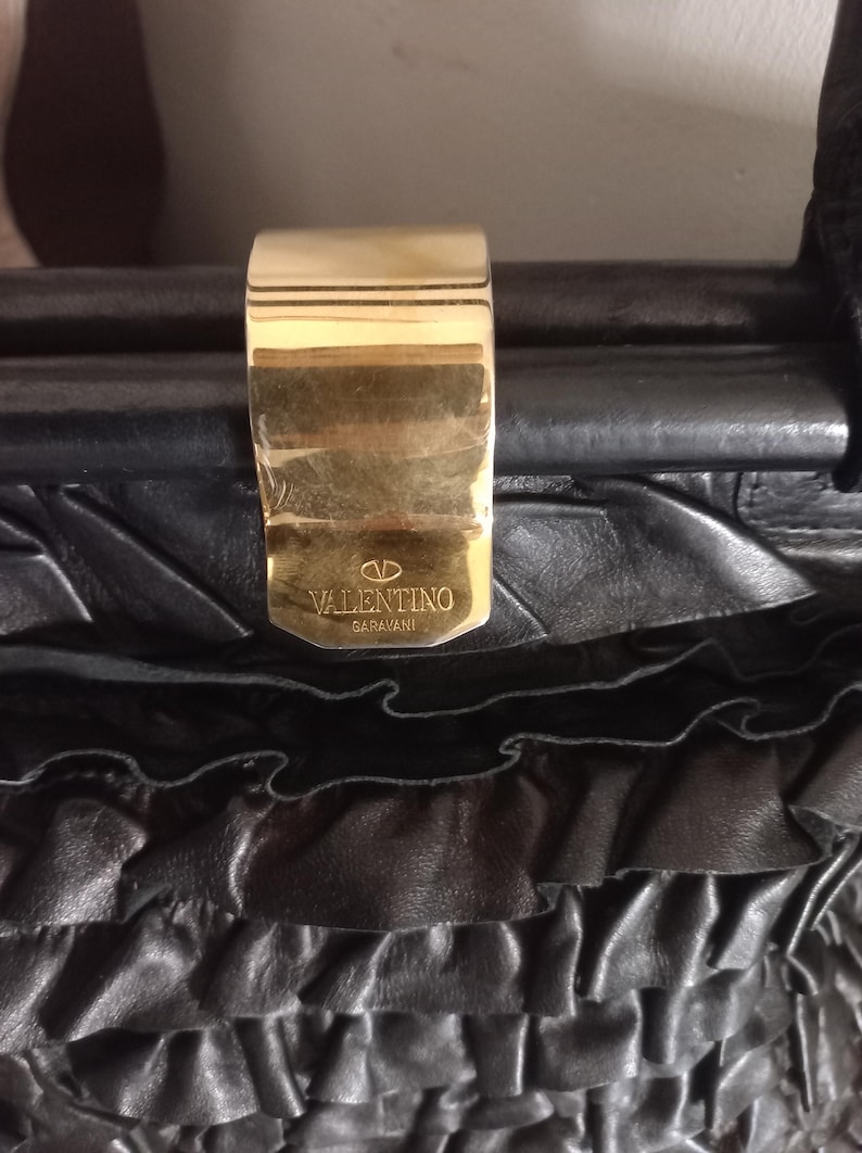 May include: Close-up of a black leather handbag with a gold clasp. The clasp is rectangular and features the Valentino Garavani logo. The bag has a textured, ruffled design.