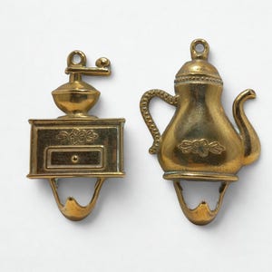 以下が含まれることがあります： 真鍮色の装飾用フック2つ。1つはコーヒーグラインダー、もう1つはティーポットの形をしています。どちらも上部に吊り下げ用のループがあり、下部に湾曲したフックが付いています。コーヒーグラインダーのフックには引き出しのディテールがあります。