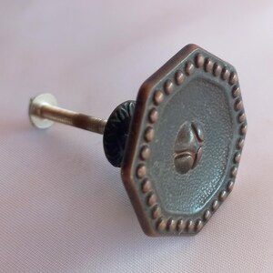May include: A hexagonal cabinet knob with a dark copper-toned finish. The face features a textured center, a raised central emblem, and a border of small, evenly spaced raised dots. A threaded metal bolt extends from the back of the knob.