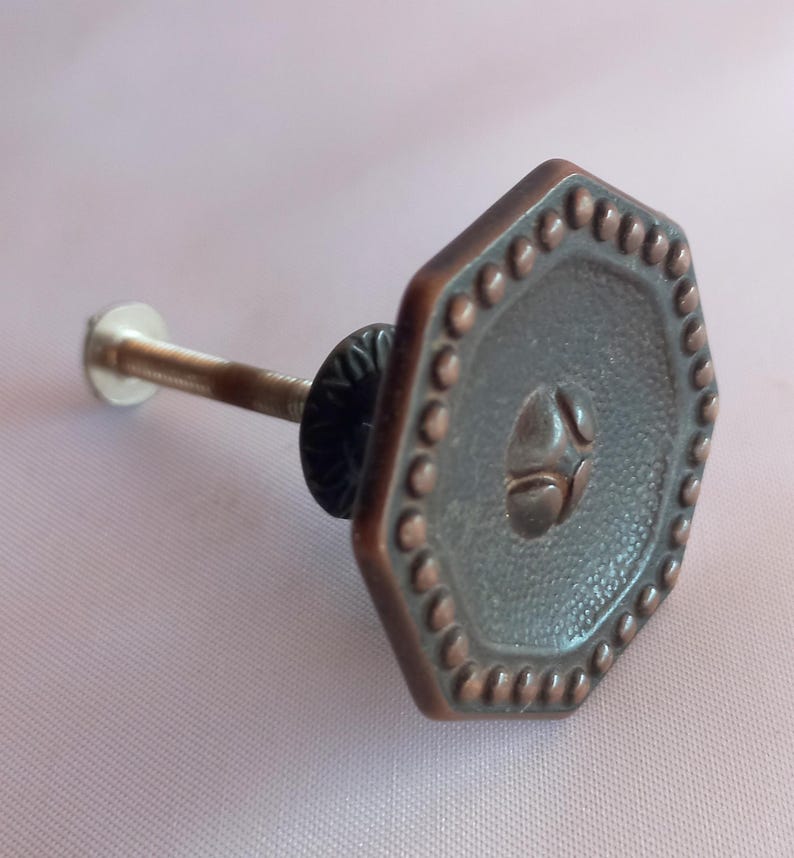 May include: A hexagonal cabinet knob with a dark copper-toned finish. The face features a textured center, a raised central emblem, and a border of small, evenly spaced raised dots. A threaded metal bolt extends from the back of the knob.