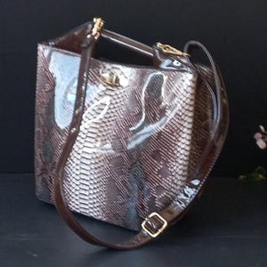 Puede incluir: Un bolso marrón y beige con estampado de piel de serpiente y acabado brillante. El bolso tiene un asa superior y una correa para el hombro desmontable. Los herrajes dorados acentúan el bolso, incluyendo el cierre y las hebillas.