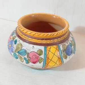 May include: A small, round ceramic pot with a wide opening. The pot is painted with floral designs in blue, pink, and green on a white background. The rim is yellow and brown, and the interior is a darker brown color.