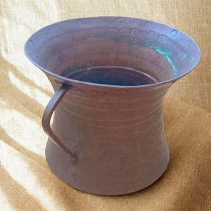 Könnte beinhalten: Ein Kupfergefäß mit weiter Öffnung und einem einzelnen geschwungenen Griff an der Seite. Die Außenfläche weist eine dunkle, verwitterte Patina mit horizontalen Bändern und einer Reihe kleiner, kreisförmiger Vertiefungen am Rand auf.