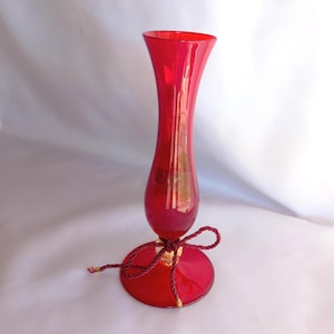 May include: A tall, ruby red glass vase with a flared top and a round base. The vase is adorned with a burgundy-colored ribbon tied around the base. The vase is approximately 10 inches tall.