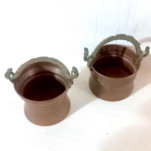 May include: Two small, copper-colored metal buckets with decorative handles. The buckets have a flared shape and a dark interior. The handles are a contrasting color, possibly bronze or a similar metal.