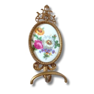 Może przedstawiać: Owalna, antyczna, złota ramka na zdjęcia z kwiatowym wzorem. Ramka zawiera porcelanową wkładkę z namalowanym bukietem różowych, białych, żółtych i fioletowych kwiatów. Ramka ma ozdobną górę i zakrzywioną podstawę.