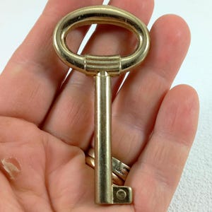 Könnte beinhalten: Ein goldfarbener Schlüssel mit einem ovalen Kopf und einem langen, geraden Schaft. Der Schlüssel wird in einer Hand gehalten.