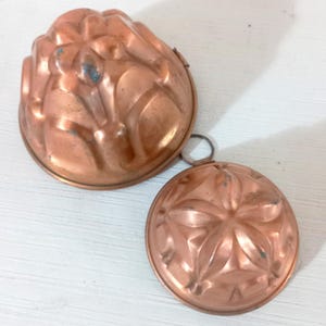 Puede incluir: Dos moldes de gelatina vintage de color cobre. Uno es un molde grande en forma de cúpula con un diseño floral. El otro es un molde redondo más pequeño con un patrón en forma de estrella. Ambos tienen un anillo para colgar.