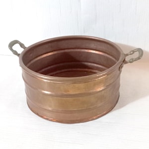 May include: A copper-colored metal bucket with two curved handles. The bucket has a cylindrical shape with horizontal bands around the exterior. The handles are a darker shade of metal and are attached to the rim of the bucket.