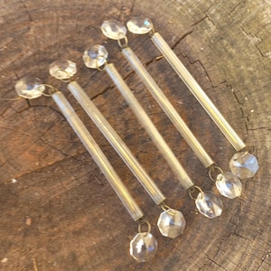 Può includere: Quattro prismi per lampadario in vetro trasparente, ciascuno composto da una lunga e sottile asta di vetro cilindrica collegata a un cristallo ottagonale sfaccettato su entrambe le estremità tramite piccoli anelli metallici, disposti in parallelo.