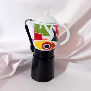 May include: A white ceramic mug with a black handle and the text "But First, Coffee" in a bold, black font. The mug is set against a blurred background, suggesting a cosy setting.