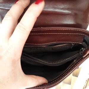 May include: Open view of a brown leather handbag, revealing the interior compartments. The bag has a flap closure and visible stitching along the edges. The interior is lined with a dark fabric. The bag is held open by a hand.