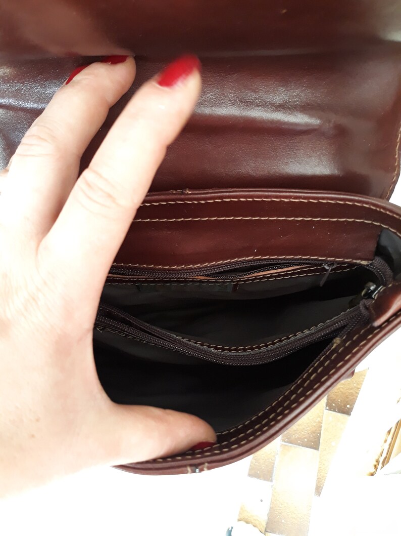 May include: Open view of a brown leather handbag, revealing the interior compartments. The bag has a flap closure and visible stitching along the edges. The interior is lined with a dark fabric. The bag is held open by a hand.
