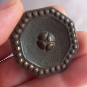 May include: An octagonal button with a dark bronze finish, featuring a textured surface, a border of small raised dots, and a central four-petaled floral motif. The button has a distinct geometric shape and a metallic appearance with subtle highlights.