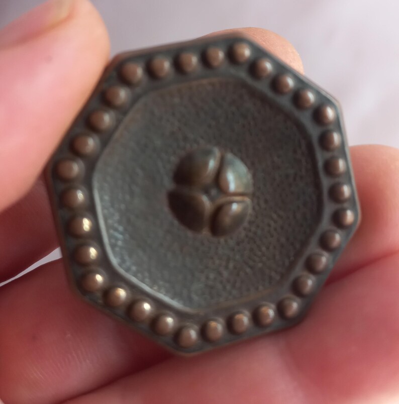 May include: An octagonal button with a dark bronze finish, featuring a textured surface, a border of small raised dots, and a central four-petaled floral motif. The button has a distinct geometric shape and a metallic appearance with subtle highlights.