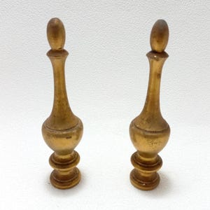 May include: Two antique gold-colored decorative finials. Each finial has a rounded top, a slender, elongated body, and a base with stacked circular elements. The objects are set against a white background.