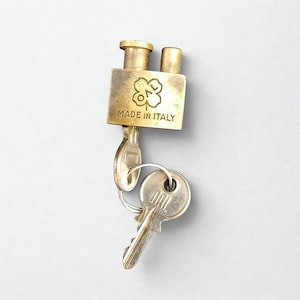 May include: A vintage brass padlock with two keys. The padlock has a rectangular body with a clover-like emblem and the text "MADE IN ITALY". The keys are attached to a metal ring.