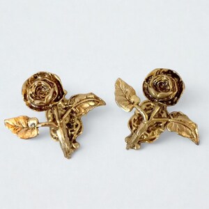 Puede incluir: Un par de pendientes dorados con forma de rosa. Cada pendiente presenta una flor de rosa detallada con varias hojas. Los pendientes están hechos de un material metálico brillante y están sobre un fondo blanco.
