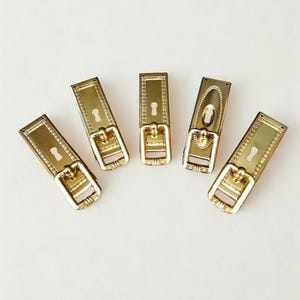 May include: Five brass-colored keyhole covers with decorative detailing. Each cover has a rectangular shape with a keyhole and a decorative pull. The covers are arranged on a white surface.