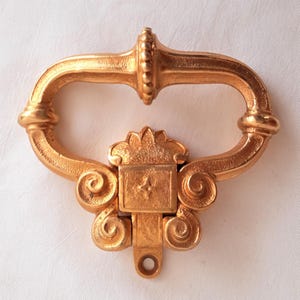 May include: A gold-colored, ornate door pull with a curved handle and decorative scrollwork. The pull features a rectangular center piece with a star design and a small hole at the bottom for mounting. The overall design is vintage.