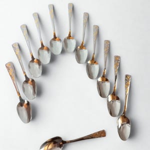 May include: A collection of silver spoons with gold-coloured bowls and handle detailing, arranged in a semi-circle against a white background. The spoons are presented in an overhead shot, showcasing their elegant design and craftsmanship.