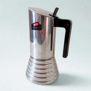 May include: A stainless steel stovetop espresso maker with a black handle and lid. The base has horizontal ridges. The brand name "VIGANO" is displayed on a red and black label on the front of the coffee pot.