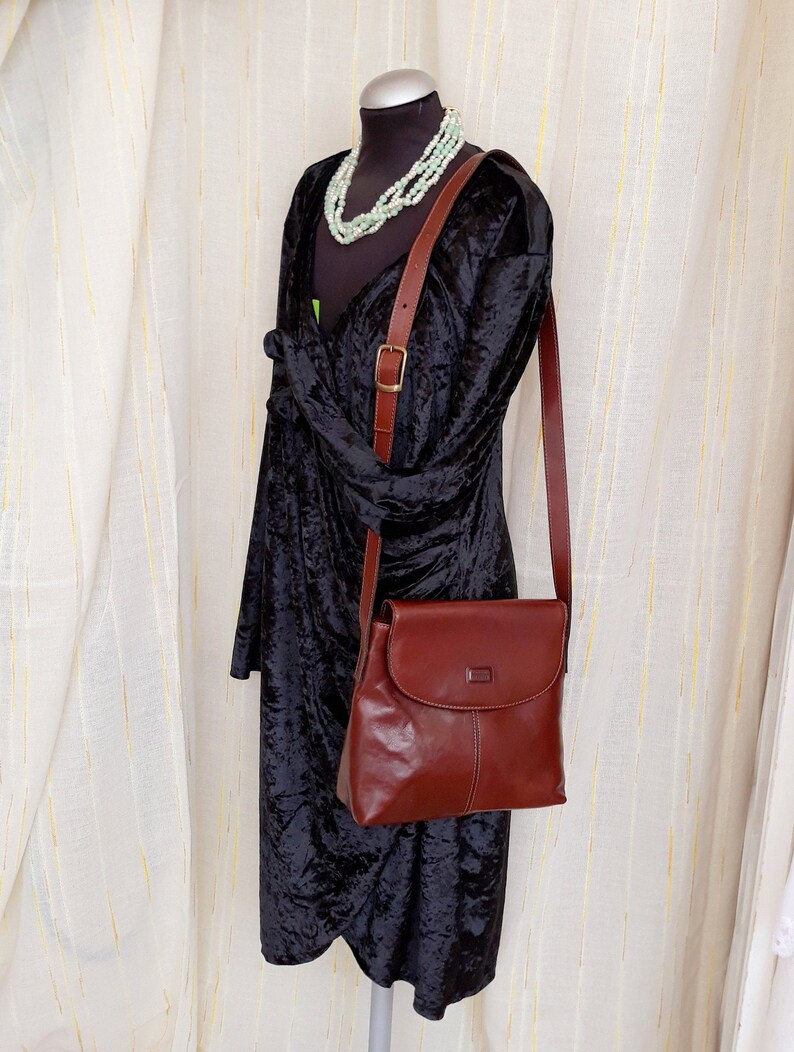 May include: A brown leather crossbody bag with a flap closure and a long adjustable strap. The bag is displayed on a mannequin wearing a black velvet dress and a multi-strand beaded necklace. The bag has a rectangular shape.