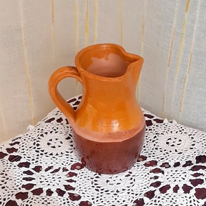 Puede incluir: Una pequeña jarra de cerámica hecha a mano, con asa y pico vertedor. La jarra es principalmente naranja con una base marrón oscuro. El interior es de color rosa claro. La jarra está sobre un mantelito blanco de encaje.