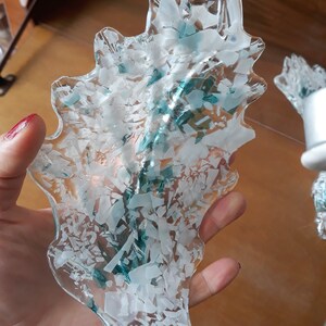 K&ouml;nnte beinhalten: Ein dekoratives St&uuml;ck aus klarem Glas in Blattform, gef&uuml;llt mit wei&szlig;en und t&uuml;rkisblauen Scherben. Das Glas wird in einer Hand gehalten, wodurch sein kompliziertes Design und seine Textur hervorgehoben werden.